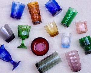 Colorful glassware laid on an off-white background. Glasses are a variety of textures. Colors include blue, grey, green, amber, pink, and red.